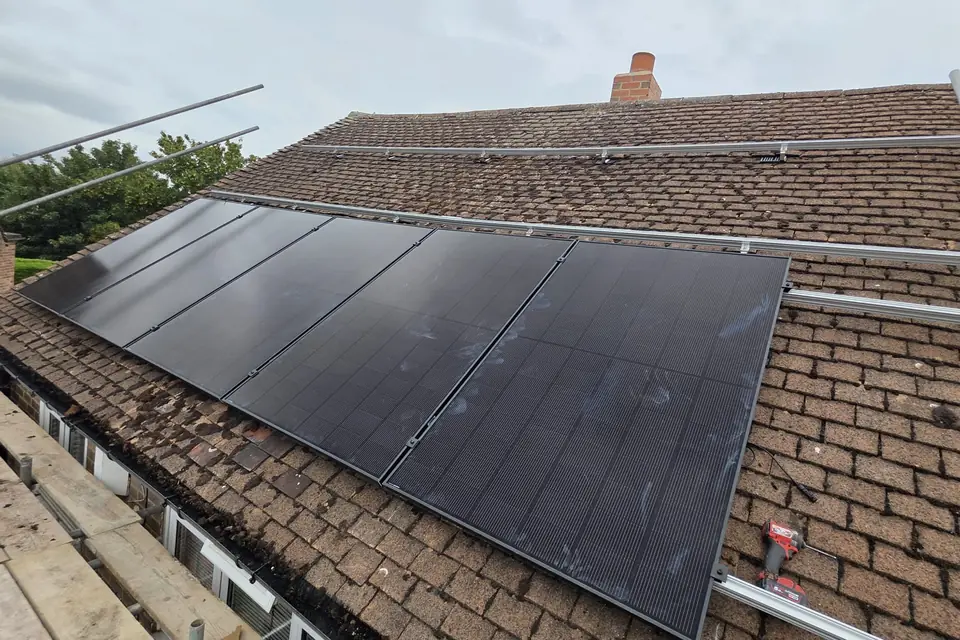 A row of black solar panels being fitted on a brown tiled roof, with empty mounting rails above it and scaffolding along the edge