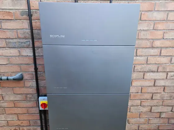 A grey home battery and its isolator switch fixed to a red brick wall above gravel