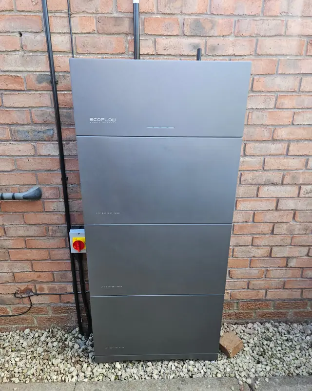 A grey home battery and its isolator switch fixed to a red brick wall above gravel