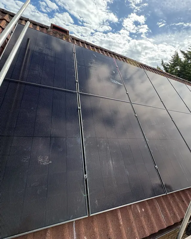 Black solar panels across a red-tiled roof under a blue sky with clouds