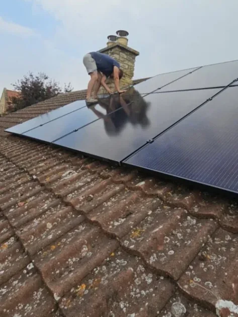 An installer fitting black solar panels on a tiled roof, beside a stone chimney