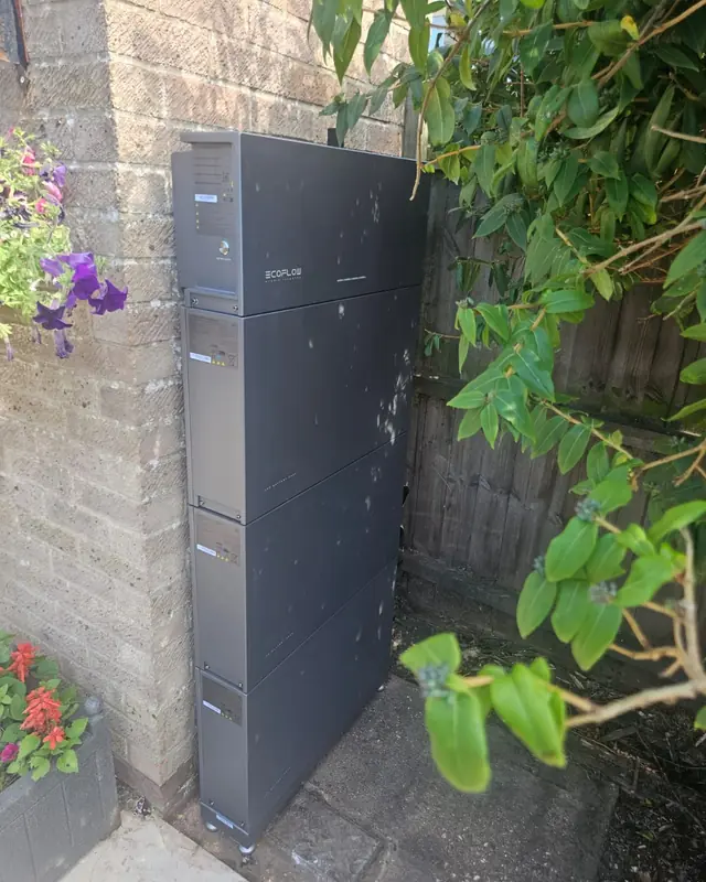 A grey home battery fixed to an outside brick wall, beside a garden fence and flowers