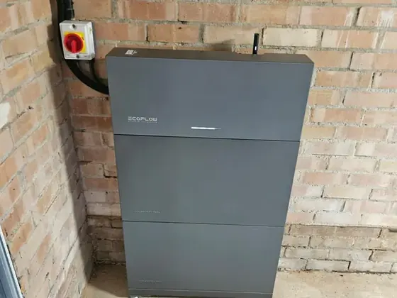 A grey home battery and its isolator switch against a brick wall in a garage