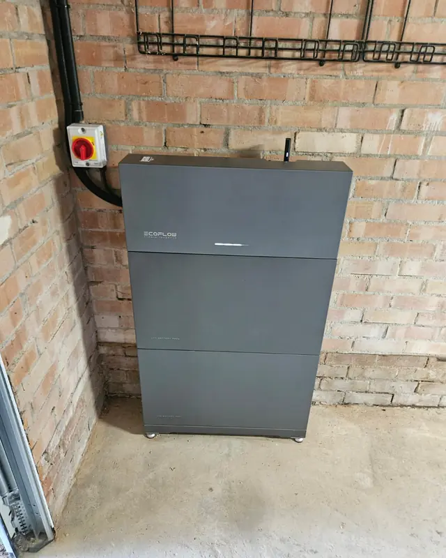 A grey home battery and its isolator switch against a brick wall in a garage