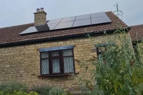 Black solar panels fitted around the chimney of a stone bungalow, with a cottage garden in front